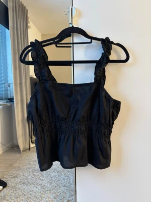 Svart linnetopp med rynkade axelband - Säljer en svart linnetopp med breda, rynkade axelband och smockad midja. Toppen har en luftig och croppad passform som ger en cool look. Perfekt att matcha med jeans eller kjol för en avslappnad stil. 