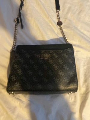 Fake Guess väska  - Snygg svart axelväska från Guess med silverfärgade kedjedetaljer och logomönster över hela väskan. Väskan har en dragkedja på baksidan och justerbar axelrem i både läder och kedja. Perfekt för dig som gillar stilrena accessoarer med en lyxig touch. Fake. 