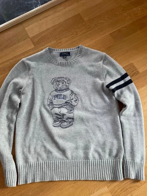 Grå stickad tröja Polo Bear - Grå stickad tröja från Polo Ralph Lauren med den ikoniska Polo Bear-figuren på bröstet. Tröjan har rund hals, ribbade muddar och två mörkblå ränder på ena ärmen. Perfekt för dig som gillar klassisk streetwear med en twist.