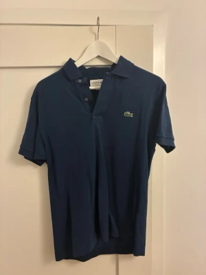 Marinblå polotopp från Lacoste - Säljer en klassisk marinblå polotopp från Lacoste med ikonisk krokodillogo på bröstet. Toppen har korta ärmar, krage och knappar framtill. Materialet är bomull och passformen är normal. Perfekt för dig som gillar stilrena och sportiga plagg.