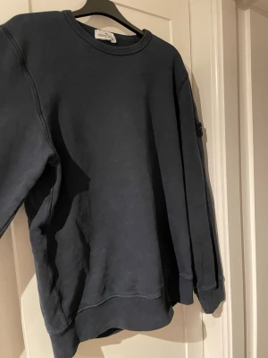 Blå sweatshirt från Stone Island - En stilren blå sweatshirt från Stone Island med klassisk rund hals och ribbade muddar. Tröjan har den ikoniska Stone Island-loggan på vänster ärm och är tillverkad i mjukt bomullsmaterial. Perfekt för en avslappnad och trendig look. 