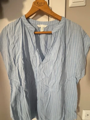 Blåvit randig blus från H&M - Ljusblå blus med vita ränder från H&M. Blusen har v-ringning, korta ärmar och är tillverkad i tunn bomull som känns luftig och skön. Perfekt till jeans eller kjol för en avslappnad look.