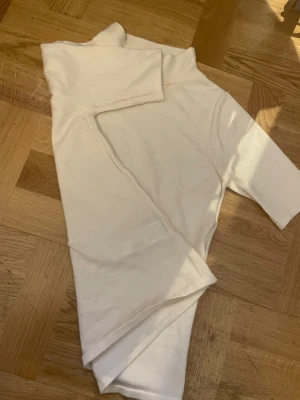 Vit t-shirt med hög krage - Säljer en vit polo t-shirt med hög krage och korta ärmar. T-shirten har en tight passform och är gjord i ett mjukt, stretchigt material som sitter snyggt på kroppen. Perfekt att styla med jeans eller kjol. Den har fina detaljer längs midjan samt vid armarna 