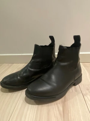 Svarta Chelsea boots i skinn - Stilrena svarta Chelsea boots i äkta skinn med klassisk rund tå och elastiska paneler på sidorna. Skorna har en låg klack och dragflikar både fram och bak för enkel påtagning. Perfekta för dig som gillar tidlös och clean stil. Dom är lite slitna men fortfarande fina!