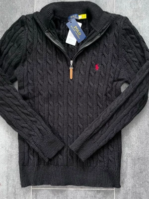 Svart Polo Ralph Lauren - Kabelstickad svart tröja från Polo Ralph Lauren med halv dragkedja och ribbade muddar. Klassisk röd logga broderad på bröstet. Tröjan har hög krage och är tillverkad i mjukt material, perfekt för lager på lager.