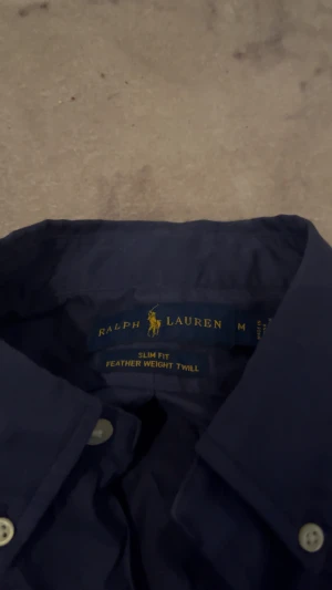 Mörkblå skjorta från Ralph Lauren - Snygg mörkblå skjorta från Ralph Lauren i slim fit med klassisk button-down krage och röda broderade loggan på bröstet. Tillverkad i lätt bomullstwill och har vita knappar längs hela framsidan. Perfekt för en clean och stilren look.