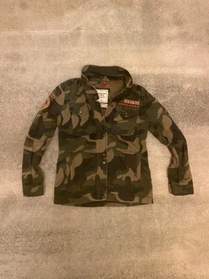 Camouflage fieldjacka från Abercrombie & Fitch  - Säljer en snygg camouflage fieldjacka från Abercrombie & Fitch. Jackan har klassisk militärgrön camomönster, flera fickor framtill, tryckknappar och coola patchdetaljer på bröstet och ärmen. Vid funderingar, fråga på! 