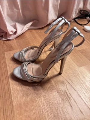Silvriga höga pumps med remmar - Snygga silvriga pumps med smala remmar över tårna och runt ankeln. Skorna har hög klack och öppen tå, vilket ger en elegant och trendig look. Perfekta för att lyfta din outfit med lite extra glans. Materialet är glansigt och insidan är mjukt vadderad för komfort.