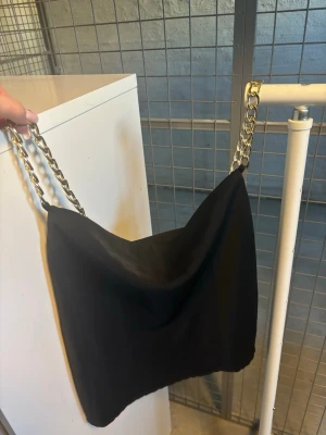 Svart bandeau tubtopp med kedjeband - Trendig svart bandeau tubtopp med breda guldfärgade kedjeband som axelband. Toppen är axelbandslös och har en enkel, stilren design som passar perfekt till många olika outfits.