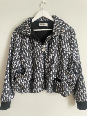 Mönstrad vindjacka från Christian Dior - Snygg vindjacka från Christian Dior med ikoniskt blått och vitt monogrammönster. Köpt som rock från Christian Dior för ca 30 000kr och insydd av en skräddare i Varberg till en vindjacka. Jackan har dragkedja framtill, huva och ribbade muddar. Två fickor med dragkedja och justerbar midja. Perfekt för dig som vill sticka ut med en exklusiv look. I storlek XL men passar perfekt på mindre storlekar också.