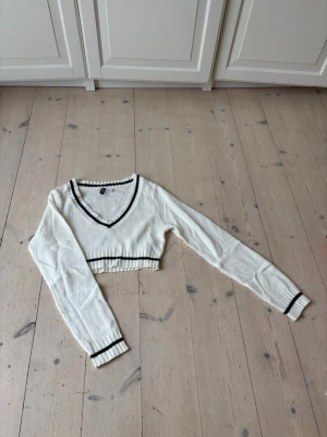 Vit croppad tröja med svarta detaljer - Säljer en vit croppad tröja med svarta ränder vid v-ringad hals, midja och ärmslut. Tröjan har långa ärmar och är stickad i ett tunt material. Perfekt för en trendig och avslappnad stil.