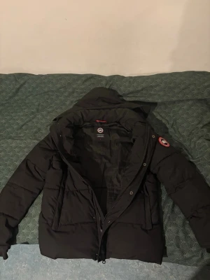 Svart dunjacka från Canada Goose - Snygg svart dunjacka från Canada Goose med klassisk logotyp på ärmen. Jackan har hög krage, dragkedja och tryckknappar framtill samt vadderad design för extra värme. Perfekt för kalla dagar och riktigt stilren look.