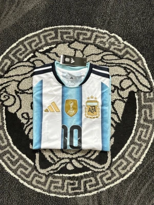 Argentina matchtröja Adidas nr 10 - Säljer en officiell Argentina matchtröja från Adidas med klassiska blå och vita ränder, guldiga detaljer och nummer 10 på bröstet. Tröjan har AFA-emblem, FIFA-märke och tre mörkblå ränder på axlarna. Perfekt för fotbollsfans!