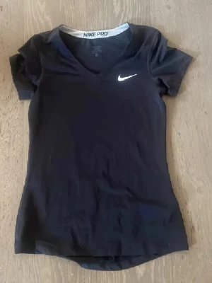 Svart Nike Pro t-shirt - Svart t-shirt från Nike Pro med vit logga på bröstet. Tröjan har en v-ringning och korta ärmar, perfekt för träning och sport. Materialet är stretchigt och andas, vilket gör den bekväm att bära under intensiva pass.