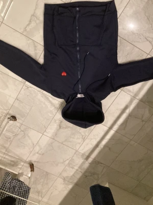 Svart hoodie från Comme des Garçons - Snygg svart hoodie med dragkedja från Comme des Garçons Play. Har det ikoniska röda hjärtat med ögon broderat på bröstet, två stora fickor fram och justerbar huva med snören. Perfekt för en chill och stilren look.