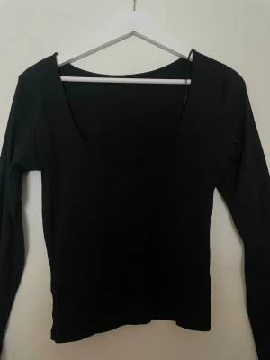 Svart långärmad topp med djup urringning - En stilren svart långärmad topp med djup urringning framtill. Toppen har en kroppsnära passform och är tillverkad i ett stretchigt, ribbat material som ger en snygg siluett. Perfekt att matcha med jeans eller kjol för en trendig look. Säljer en i blå 