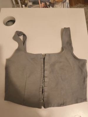 Grå croppad linnetopp med dragkedja - Snygg grå croppad linnetopp med breda axelband och dragkedja framtill. Toppen har en enkel och stilren design som passar perfekt till jeans eller kjol. Materialet känns mjukt och stretchigt för en bekväm passform.