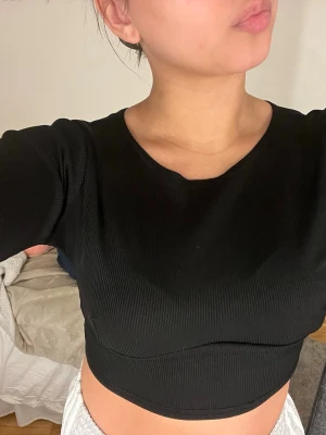 Svart croppad topp från Boohoo - Snygg svart croppad topp från Boohoo med lång ärm och rund halsringning. Toppen är ribbad och har en figurnära passform som framhäver midjan. Perfekt att matcha med höga jeans eller kjol för en trendig look.