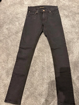 Svarta skinny jeans från Nudie Jeans - Säljer ett par svarta skinny jeans från Nudie Jeans med klassisk femficksmodell och smal passform. Jeansen är tillverkade i ekologisk bomull och har diskreta sömmar samt läderpatch bak i midjan. Perfekta för en clean och stilren look.
