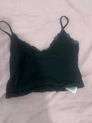 Svart spetsbody med tunna axelband - Snygg svart body med tunna axelband och spetsdetaljer längs urringningen. Mjuk och stretchig känsla, perfekt för lager på lager eller som ett statement-plagg. Enkel och stilren design som passar till mycket.