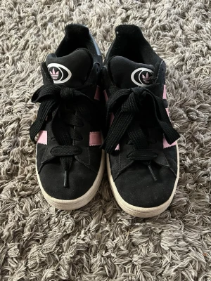 Svarta Adidas Campus med rosa ränder - Snygga Adidas Campus sneakers i svart mocka med klassiska rosa ränder på sidorna och vit sula. Tjocka svarta skosnören och stor Adidas-logga på plösen. Perfekta för dig som gillar retrostil och streetwear.