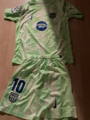 Barcelona fotbollskit limegrön Nike - Säljer ett limegrönt Barcelona fotbollskit från Nike med matchtröja och shorts. Tröjan har klubbmärke, sponsorlogga och Champions League-märke på ärmen. Shortsen har nummer 10 och klubbmärke. Perfekt för dig som älskar Barca och vill sticka ut på planen.