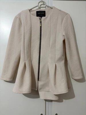 Beige kavaj från River Island - Stilren beige kavaj från River Island med rund halsringning och markerade sömmar. Kavajen har en rak passform, dragkedja framtill och lätt strukturerat tyg. 