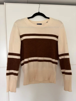 Stickad tröja från Gant  - Stickad tröja från GANT i beige med breda bruna ränder över bröstet och ärmarna samt rund halsringning. Använd några gånger och har tyvärr fått en fläck på höger arm (se bild 4). 