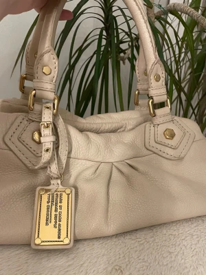Beige handväska från Marc by Marc Jacobs - Snygg beige handväska i mjukt läder från Marc by Marc Jacobs. Väskan har guldiga metalldetaljer, dubbla handtag och ett coolt innerfoder med svartvitt textmönster. Perfekt för dig som gillar stilrena accessoarer med lyxig känsla.