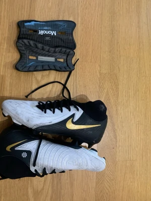 Nike fotbollsskor svart/vit/guld - Säljer ett par snygga Nike fotbollsskor i svart och vitt med guldfärgad swoosh. Skorna har klassisk snörning och dobbar. Även ett par MONOLIT benskydd som är lätta och sitter skönt. 