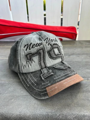Grå keps med New York 79 brodyr - Snygg grå keps med broderad text 'New York' och siffran 79 framtill. Kepsen har vita kontrastsömmar och en brun detalj på skärmen. Tillverkad i bomullstyg med en avslappnad, vintage look. Perfekt för dig som gillar streetwear och sportig stil.