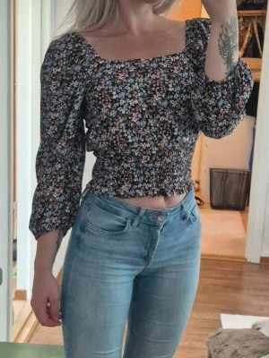 Blommig croppad blus med puffärm - Supersöt blommig blus med croppad passform och smockad midja. Toppen har trekvartsärmar med puff och fyrkantig urringning. Perfekt för dig som gillar romantisk stil och vill ha något som sticker ut lite extra.
