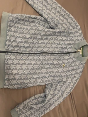 Mönstrad blå ziptröja från Lacoste - Snygg blå ziptröja från Lacoste med geometriskt mönster i vitt och ljusblått. Tröjan har ribbade muddar, dragkedja framtill och den klassiska krokodilloggan på bröstet. Perfekt för en trendig och avslappnad stil. Obs kvitto finns.