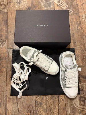 Numeris vita sneakers med camomönster - Snygga vita sneakers från Numeris med grått camomönster på sidorna. Skorna har tjocka vita snören, vit sula och detaljer i textil och mockaimitation. Perfekta för dig som vill ha en clean men ändå unik look.