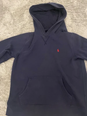 Mörkblå hoodie från Ralph Lauren - Mörkblå hoodie från Ralph Lauren med klassisk röd broderad logga på bröstet. Hoodien har stor huva och en rymlig magficka framtill. Tillverkad i mjukt bomullsmaterial som är skönt att ha på sig. Perfekt för en avslappnad och stilren look.
