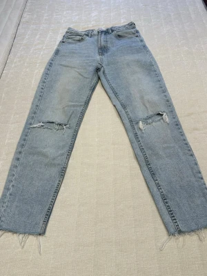 Ljusblå slitna raka jeans - Säljer ett par ljusblå jeans med raka ben och slitningar vid knäna. Jeansen har fransiga benslut och klassisk femficksmodell. Perfekta för en avslappnad och trendig look. Materialet är denim i bomull.