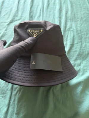  bucket hatPrada - Snygg svart bucket hat det ikoniska metalltriangelloggan framtill. Hatten är tillverkad i ett glansigt, vattenavvisande material och har en klassisk form med bred, nedåtlutande brätte. Perfekt accessoar för en trendig streetwear-look.