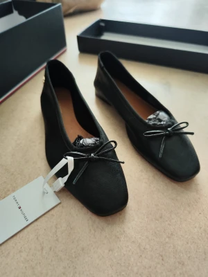 Svarta ballerinaskor Tommy Hilfiger - Stilrena svarta ballerinaskor från Tommy Hilfiger med diskret logga i guld på hälen och dekorativ rosett framtill. Skorna har en klassisk rundad tå och är tillverkade i ett slätt material. Perfekta för en trendig och enkel look.