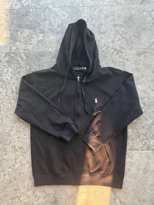 Svart hoodie från Polo Ralph Lauren - Sitter som L! Klassisk svart hoodie från Polo Ralph Lauren med dragkedja framtill och vit broderad logga på bröstet. Hoodien har huva med dragsko och känguruficka. Tillverkad i mjuk bomullsblandning som är skön att bära. Perfekt för en avslappnad och stilren look.
