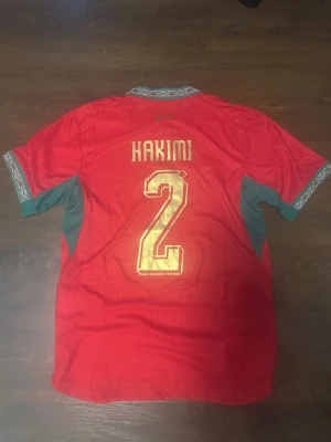 Marocko fotbollströja Hakimi #2 Puma - Röd matchtröja (player-version) från Puma med Marockos landslagsemblem och Hakimi nummer 2 i guld. Tröjan har gröna detaljer på sidorna och ärmarna samt mönstrad krage och ärmslut. Perfekt för dig som vill ha en snygg marocko tröja nu under slutspelet