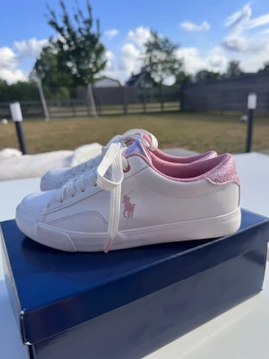 Vita och rosa sneakers från Polo Ralph Lauren - Snygga vita sneakers från Polo Ralph Lauren med rosa detaljer och logga på sidan. Skorna har vita snören, rosa insida och glitterdetalj på hälen. Tillverkade i syntetmaterial och textil för en fräsch look. Använda 2 gånger 