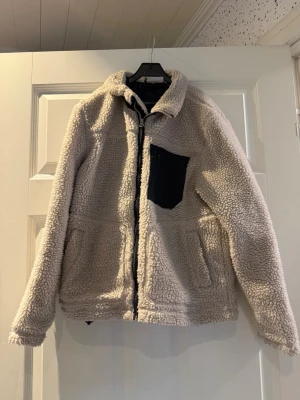 Beige teddyjacka med svart ficka - Mysig beige teddyjacka med dragkedja framtill och två sidofickor. Jackan har en svart bröstficka som kontrast och en hög krage för extra värme. Perfekt för kyliga dagar och ger en avslappnad, trendig look. Materialet är fluffigt och mjukt, typiskt för teddy. Strl 154-158