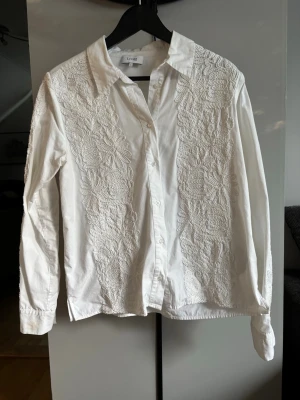 Vit skjorta/blus från Leveté Room - Stilren vit skjorta från Levete med långa ärmar och knappar framtill. Skjortan har stora broderade blommor på framsidan och på armarna som ger en unik och trendig look. Perfekt för dig som gillar detaljer och vill sticka ut lite extra.