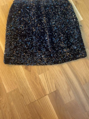 Svart paljettkjol från Gina Tricot - Säljer en svart kortkjol från Gina Tricot täckt med glittriga paljetter i svart och silver. Kjolen har en rak passform och är perfekt för dig som vill sticka ut med lite extra bling på festen. Fodrad insida för bekväm känsla. Har endast testat kjolen tvättas innan jag skickar den 