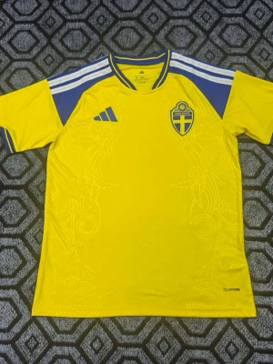 Svensk landslagströja Adidas Hemma - Gul och blå fotbollströja från Adidas med Sveriges landslagsemblem på bröstet och vita ränder på axlarna. Tillverkad i lätt och ventilerande funktionsmaterial, perfekt för träning eller match. Diskreta mönster i tyget och 'Sverige' tryckt i nacken.