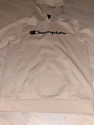 Beige hoodie från Champion - Snygg beige hoodie från Champion med broderad logga på bröstet och liten logga på ärmen. Klassisk känguruficka framtill och huva. Perfekt för en avslappnad streetwear-look. Mjuk bomullsblandning som är skön att ha på sig.