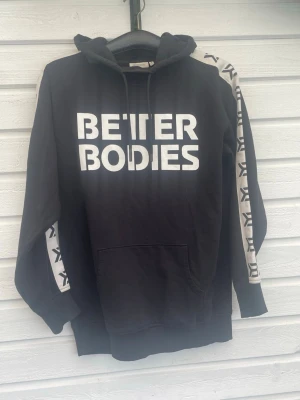 Svart Better Bodies hoodie - Svart hoodie från Better Bodies med stor vit logga på bröstet och vita ränder med svart mönster längs ärmarna. Klassisk huva med dragsko och stor magficka framtill. Tillverkad i mjukt bomullsmaterial, perfekt för en sportig och avslappnad stil.