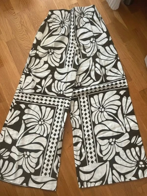 Svarta och vita linnebyxor med blommönster - Säljer ett par vida linnebyxor i svart och vitt med stort blommigt mönster och grafiska detaljer. Byxorna har resår i midjan och är tillverkade i ett lätt och luftigt linnetyg. Perfekta för en avslappnad och trendig stil.