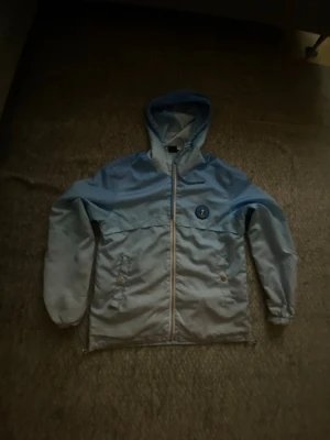Ljusblå Trapstar Windbreaker  - Snygg ljusblå vindjacka med huva och dragkedja framtill. Jackan har två fickor med knappar och ett tryckt märke på bröstet. På ryggen finns ett tryck i mörkare blått. Perfekt för blåsiga dagar och enkel att matcha till olika outfits. 