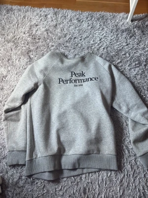 Grå sweatshirt Peak Performance - Säljer en stilren grå sweatshirt från Peak Performance med svart logga och text på bröstet. Tröjan har rund halsringning, ribbade muddar och mjukt fleecefoder på insidan. Perfekt för en avslappnad och sportig look.